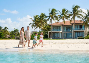Une famille heureuse marche sur une plage de sable blanc devant une villa de luxe entourée de palmiers sous un ciel bleu.