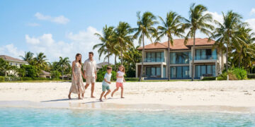 Une famille heureuse marche sur une plage de sable blanc devant une villa de luxe entourée de palmiers sous un ciel bleu.