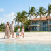 Une famille heureuse marche sur une plage de sable blanc devant une villa de luxe entourée de palmiers sous un ciel bleu.