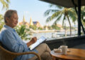 Une personne âgée utilisant une tablette sur une terrasse avec vue sur un temple thaïlandais et une rivière.