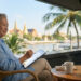Une personne âgée utilisant une tablette sur une terrasse avec vue sur un temple thaïlandais et une rivière.