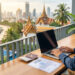 Un freelance travaillant sur son ordinateur avec un permis de travail thaïlandais et un passeport devant une vue sur la mer.