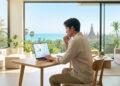 Un homme travaillant sur un ordinateur portable dans un salon moderne avec vue sur la mer et un temple en arrière-plan.
