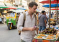 Un touriste tenant des billets thaïlandais devant un stand de nourriture de rue en Thaïlande avec un tuk-tuk en arrière-plan.