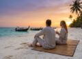 Couple assis sur une plage en Thaïlande au coucher du soleil, regardant des bateaux traditionnels sur l'eau.