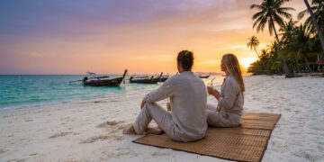 Couple assis sur une plage en Thaïlande au coucher du soleil, regardant des bateaux traditionnels sur l'eau.