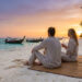Couple assis sur une plage en Thaïlande au coucher du soleil, regardant des bateaux traditionnels sur l'eau.