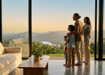 Une famille regarde par une baie vitrée un paysage vallonné au coucher du soleil dans un salon moderne.