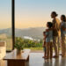 Une famille regarde par une baie vitrée un paysage vallonné au coucher du soleil dans un salon moderne.
