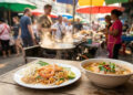 Assiette de Pad Thaï et bol de soupe traditionnelle posés sur une table en bois dans un marché de rue animé en Thaïlande.