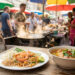 Assiette de Pad Thaï et bol de soupe traditionnelle posés sur une table en bois dans un marché de rue animé en Thaïlande.