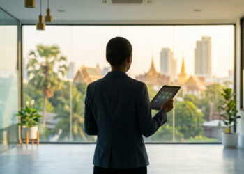 Une femme d'affaires regarde par la fenêtre d'un bureau moderne avec vue sur Bangkok et tient une tablette numérique.