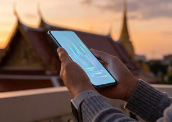 Mains tenant un smartphone avec des graphiques lumineux sur l'écran, avec un temple thaïlandais en arrière-plan au coucher.