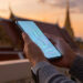 Mains tenant un smartphone avec des graphiques lumineux sur l'écran, avec un temple thaïlandais en arrière-plan au coucher.