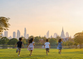 Quatre enfants courant joyeusement sur une pelouse verte face à l'horizon de Bangkok et ses temples au crépuscule.