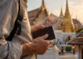 Un voyageur tient un passeport et une liasse de bahts thaïlandais devant un temple doré en Thaïlande.