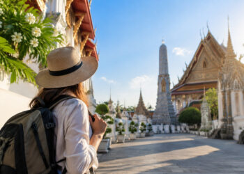 Une voyageuse avec un chapeau et un sac à dos regarde un temple bouddhiste traditionnel en Thaïlande.