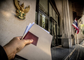 Une main tient des dossiers devant l'entrée d'un bâtiment officiel en Thaïlande avec le symbole du Garuda au mur.