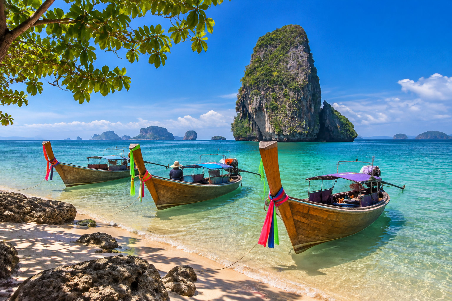 Trois bateaux traditionnels sont amarrés près d’une plage bordée de rochers, face à une grande île rocheuse couverte de végétation.