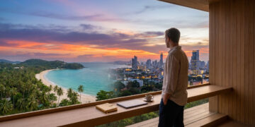 Un homme regarde un coucher de soleil sur une plage tropicale et une ville depuis un bureau moderne avec ordinateur.