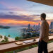 Un homme regarde un coucher de soleil sur une plage tropicale et une ville depuis un bureau moderne avec ordinateur.