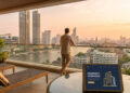 Un homme sur un balcon surplombant Bangkok avec une tablette affichant une application immobilière au premier plan.
